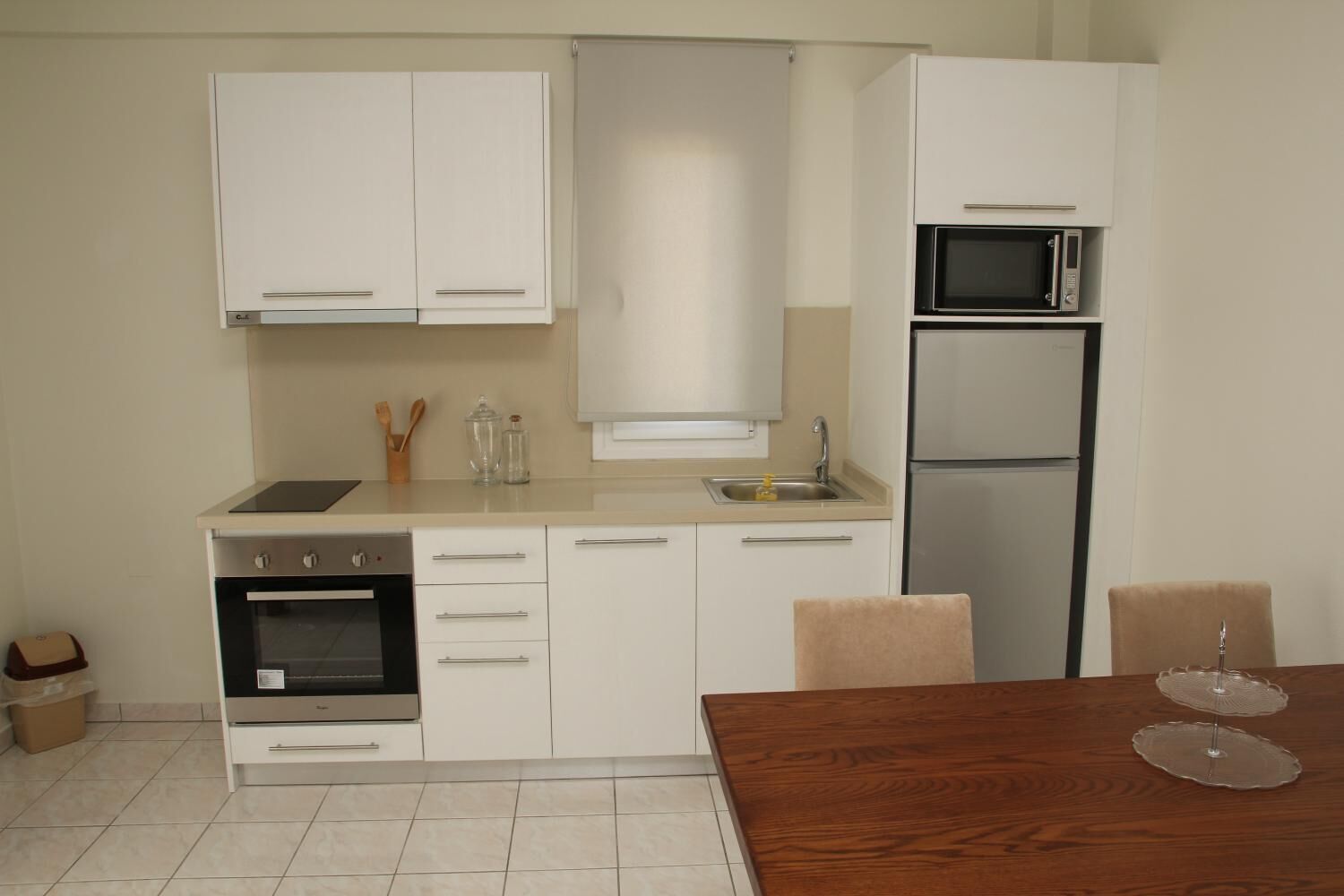 Eine kompakte Küche mit weißen Schränken, einem Backofen, einer Mikrowelle und einem Kühlschrank. Ein Holztisch mit zwei klaren Glaskuchenständern steht daneben.