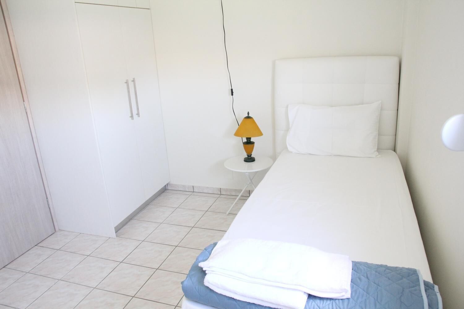 A single-bed room with a white bed, a small bedside table with a lamp, and a wardrobe.