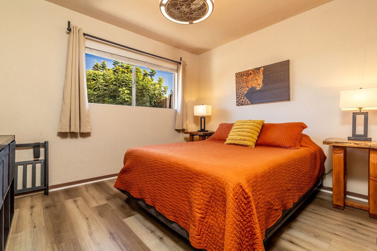 Un dormitorio con cama, ventana con cortinas, mesita de noche, lámpara y un cuadro en la pared.
