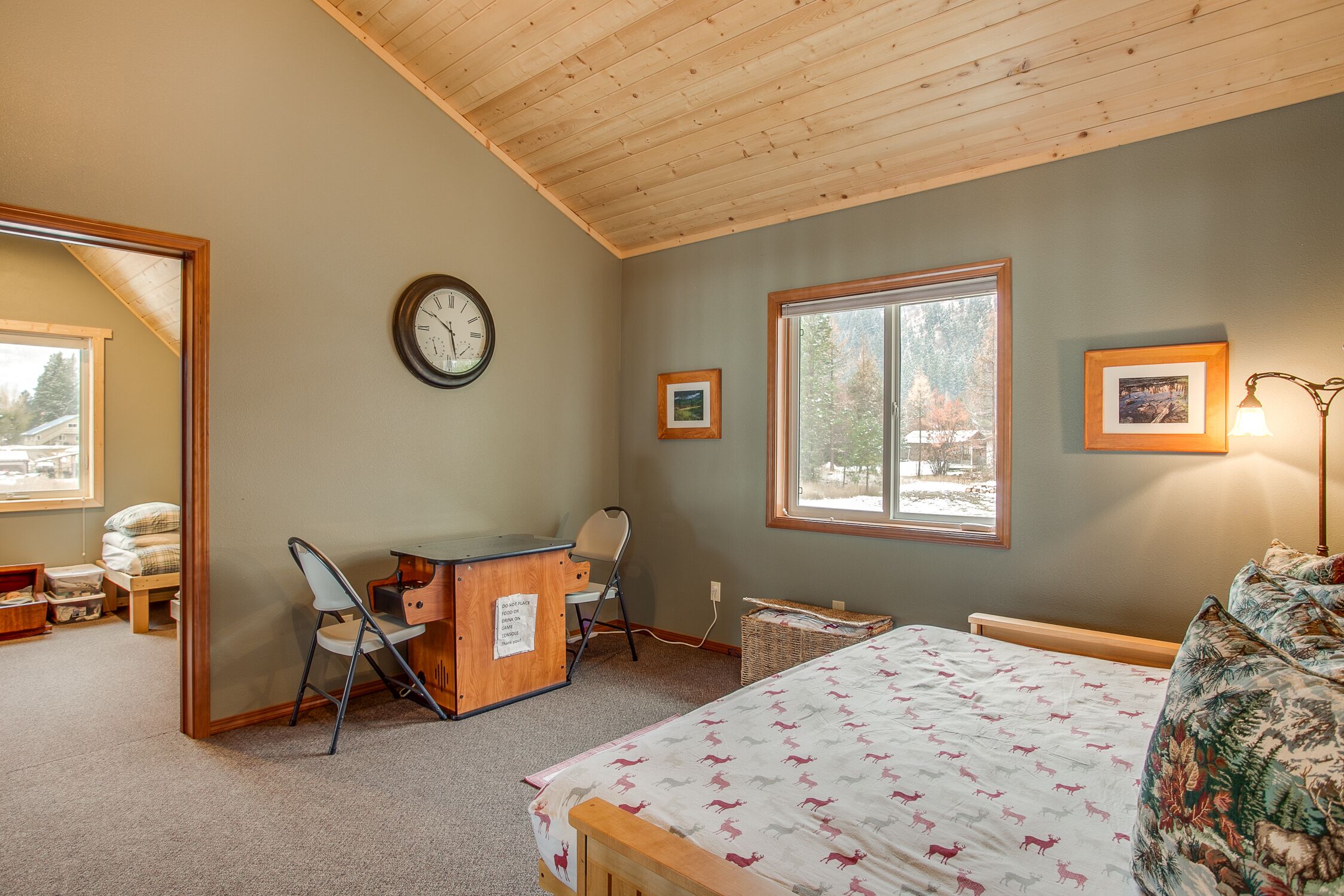 Une chambre avec un plafond en bois, un lit recouvert d’une couvre-lit à motifs, un bureau avec une chaise, une horloge et deux tableaux encadrés accrochés au mur.