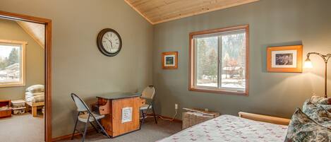 Une chambre avec un plafond en bois, un lit recouvert d’une couvre-lit à motifs, un bureau avec une chaise, une horloge et deux tableaux encadrés accrochés au mur.