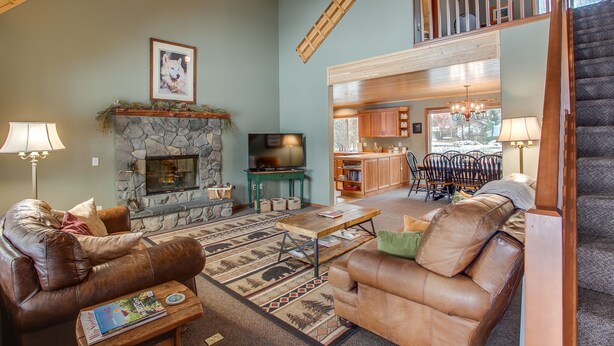 A spacious living room with a stone fireplace, leather sofas, and a wooden coffee table. A television is mounted above the fireplace, and there is a staircase leading to an upper level.
