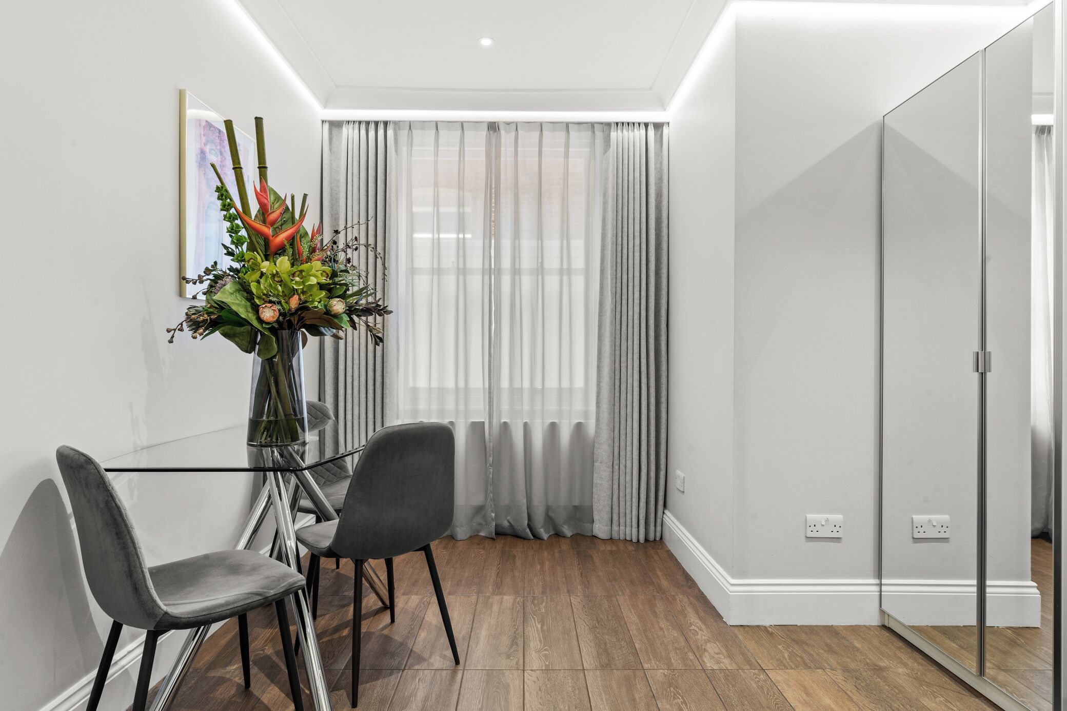 A modern room with a glass dining table, two chairs, a vase with flowers, a large window with sheer curtains, and a mirrored closet.