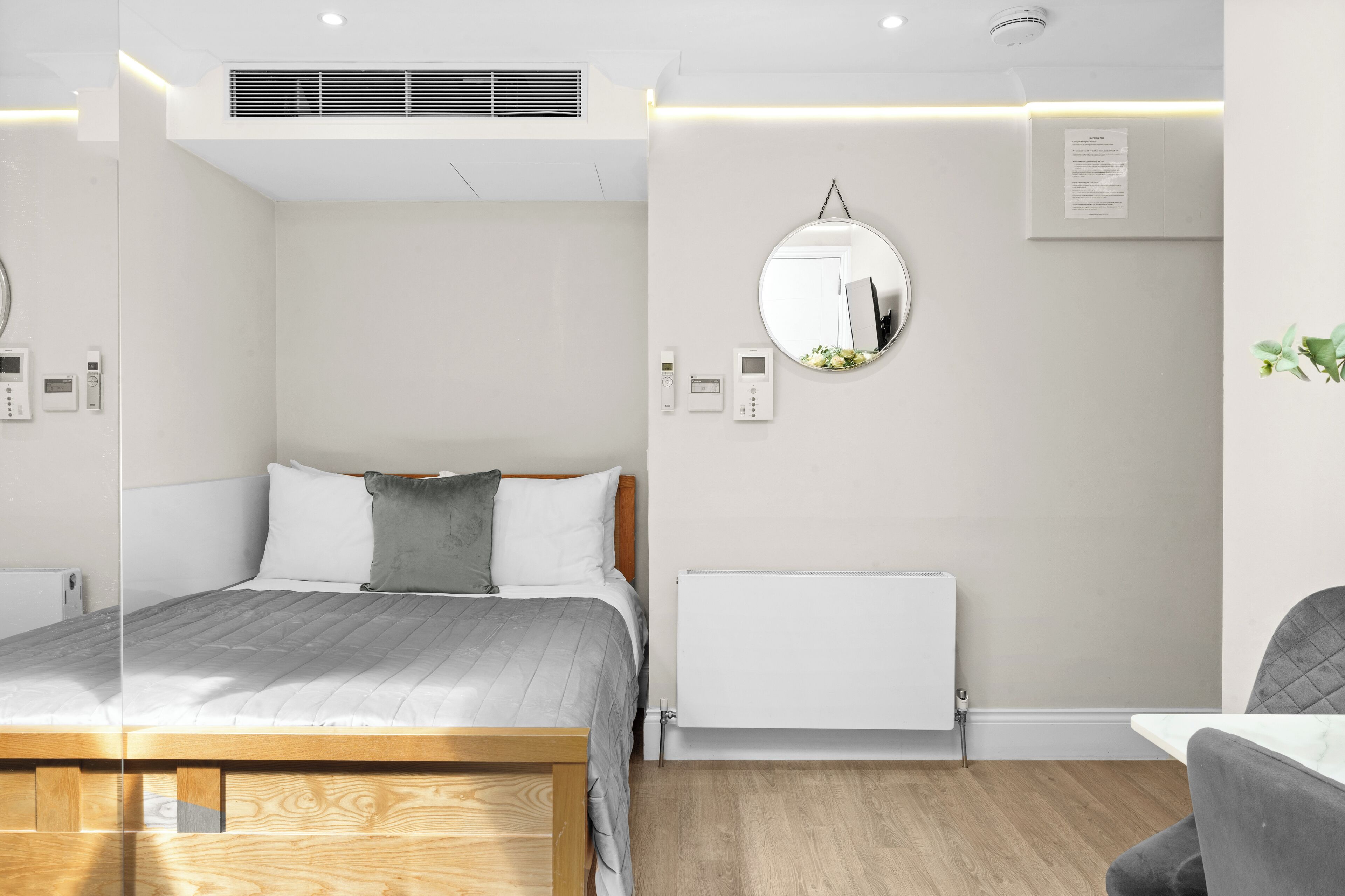 Quarto de hotel moderno com uma cama de madeira, um espelho redondo, um radiador branco e uma pequena mesa com uma cadeira.