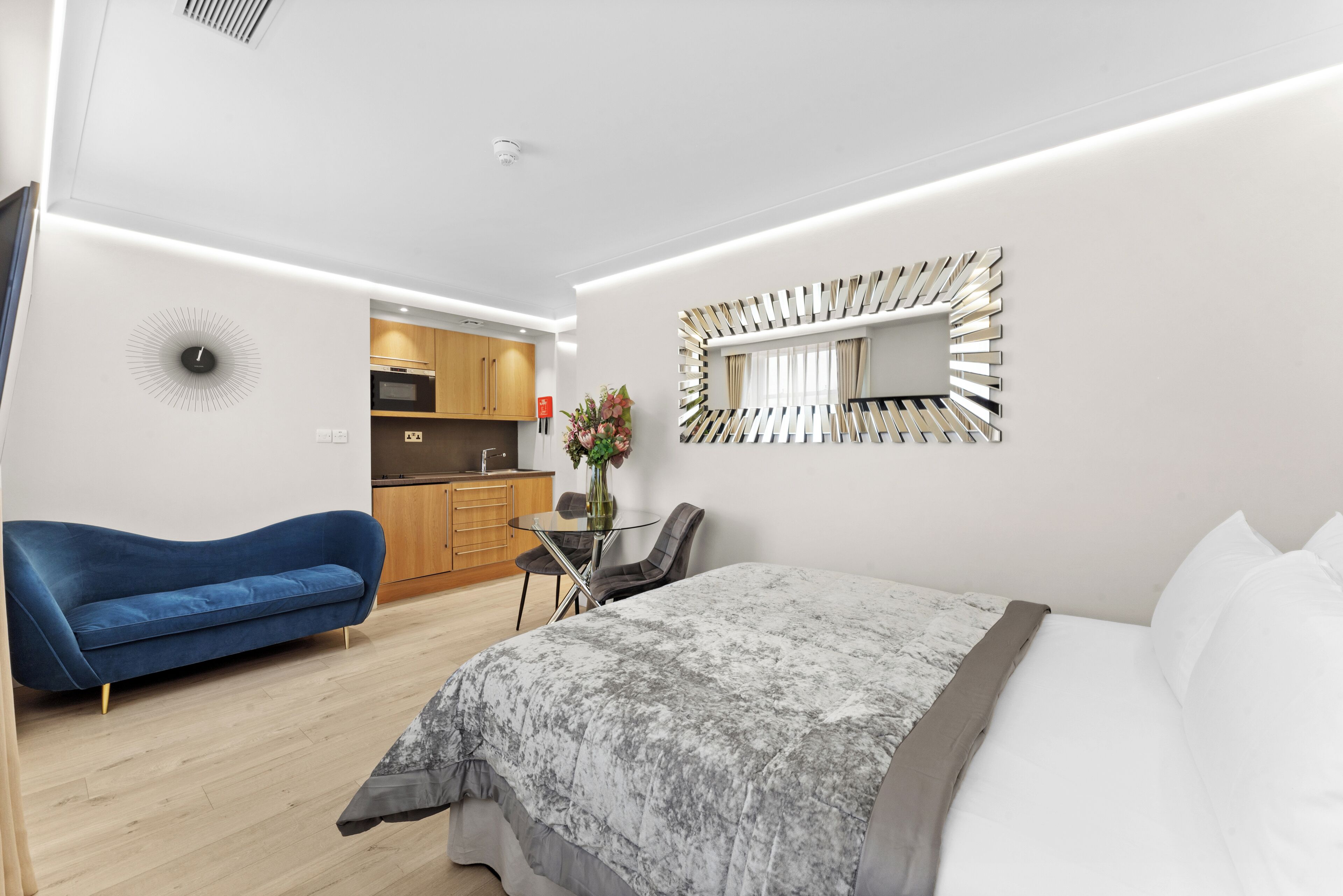 A modern hotel room with a bed, a blue sofa, a small dining table with chairs, and a kitchen area in the background.