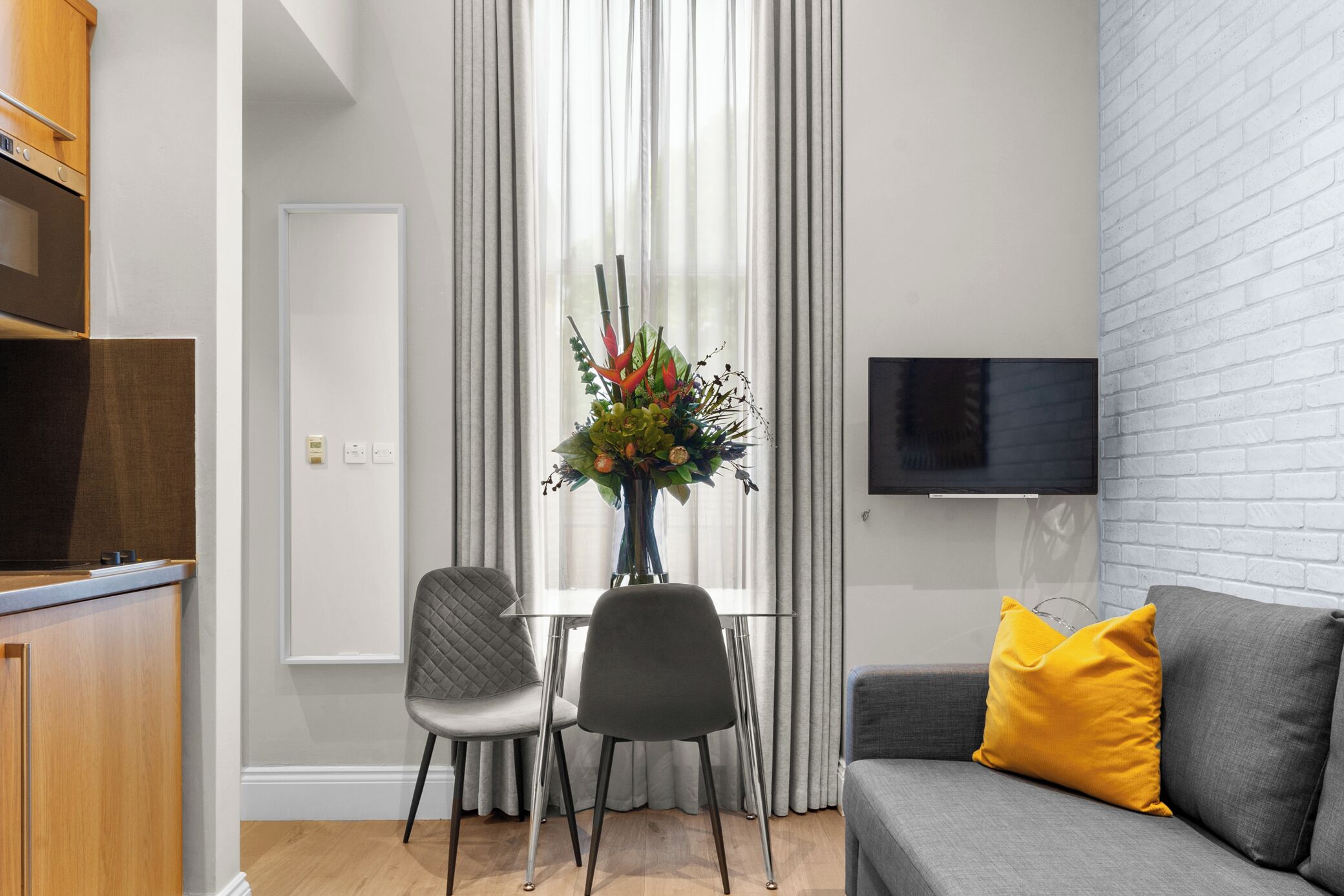 A modern living room with a grey sofa, a yellow pillow, a dining table with two chairs, a vase with flowers, and a TV mounted on the wall.