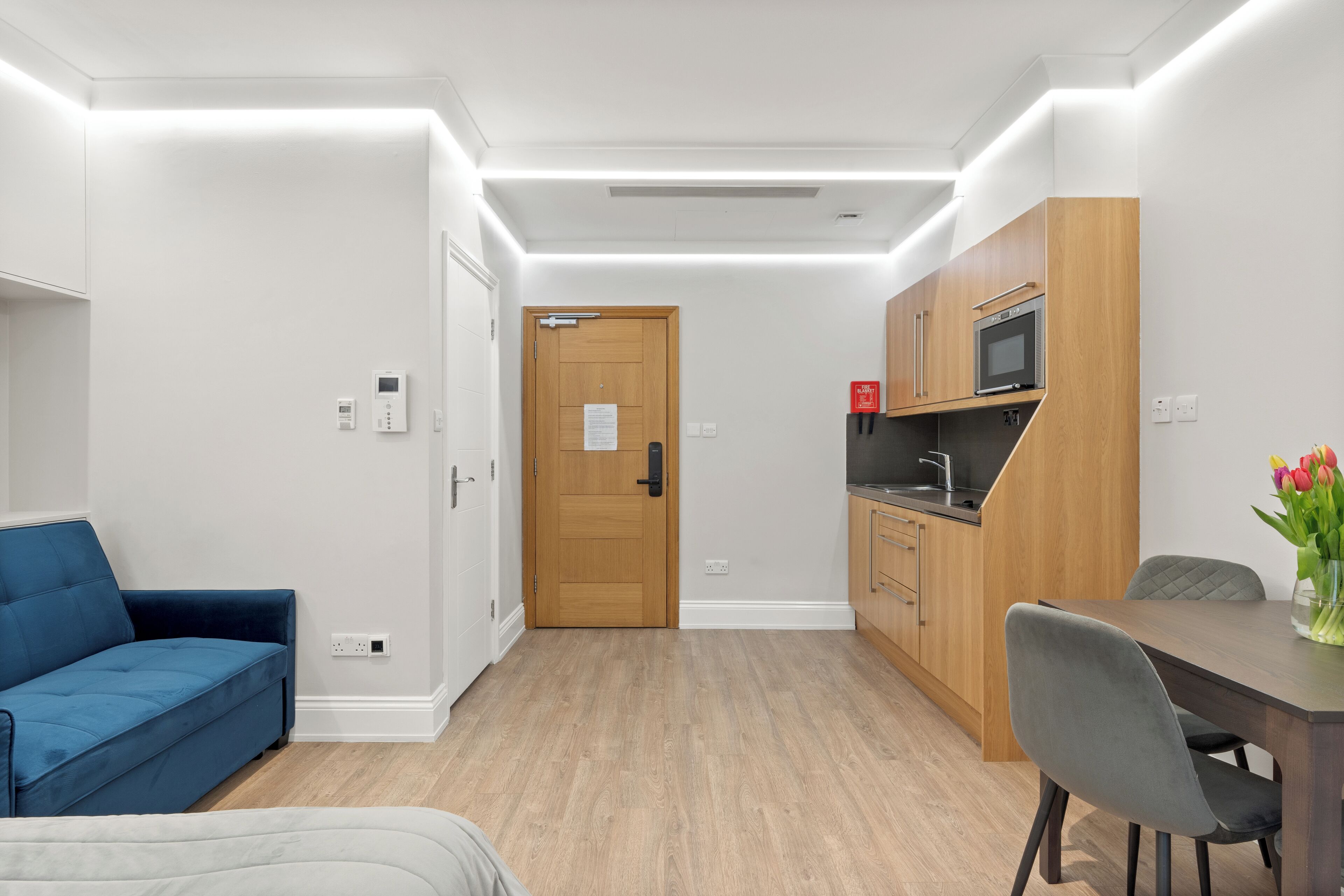 A modern hotel room with a blue sofa, a wooden dining table with two chairs, a kitchenette with a microwave, and a wooden door with a sign.