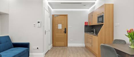 Una habitación de hotel moderna con un sofá azul, una mesa de comedor de madera con dos sillas, una kitchenette con microondas y una puerta de madera con un cartel.