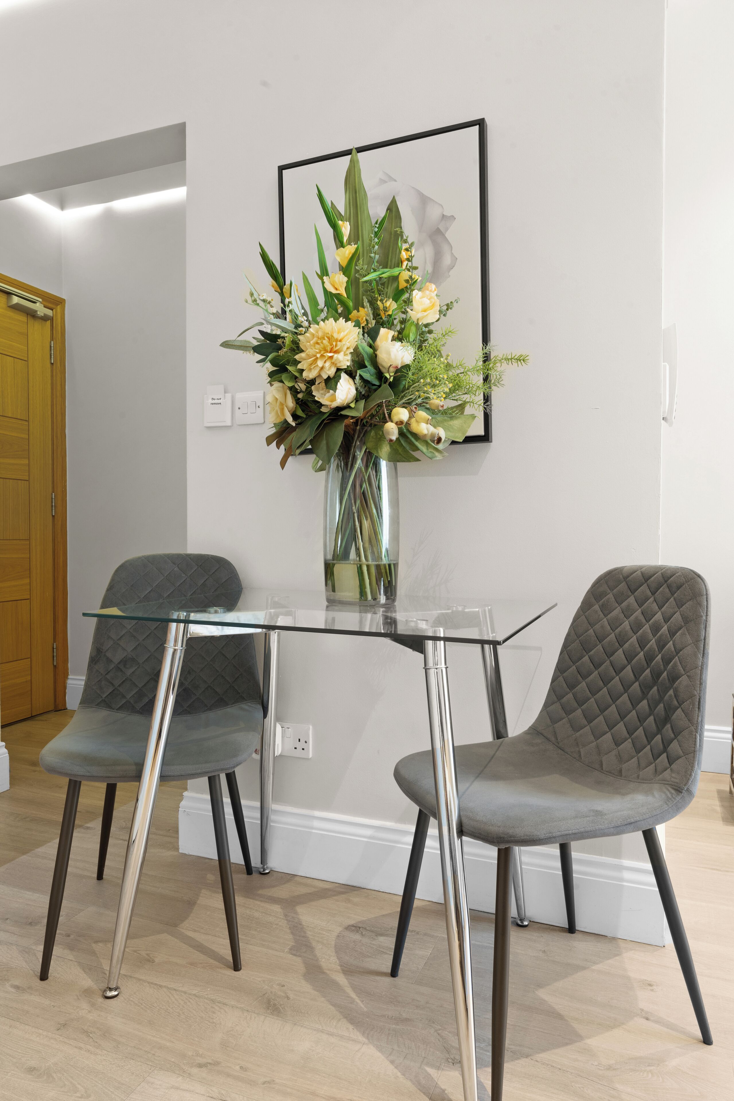 A modern room with a glass table, two grey upholstered chairs, and a vase with flowers on the wall.