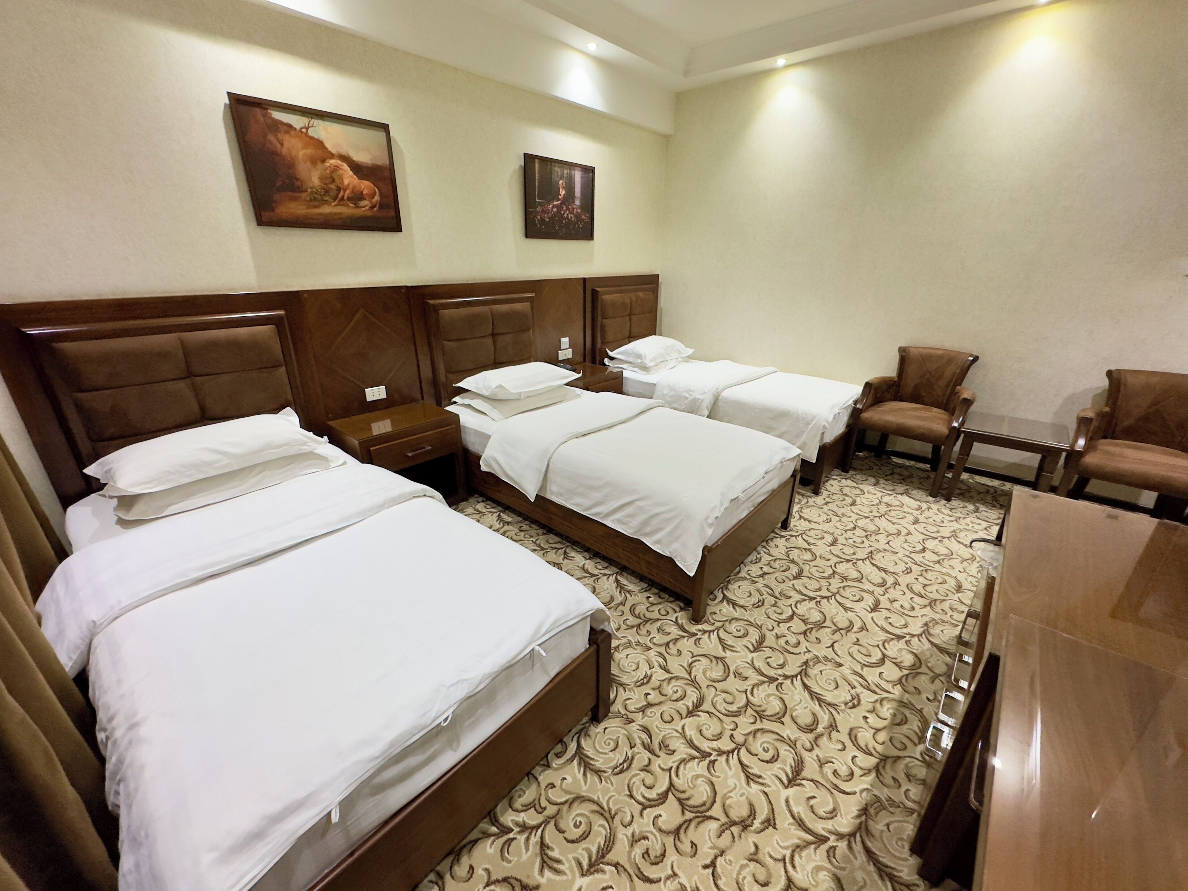 A hotel room with four beds, two nightstands, a wooden headboard, a patterned carpet, and two framed pictures on the wall.