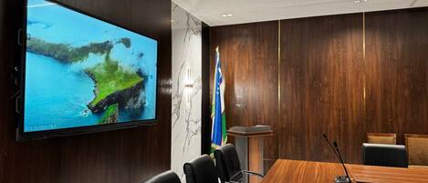 A conference room with a large flat-screen TV, wooden paneling, a podium, and black leather chairs.