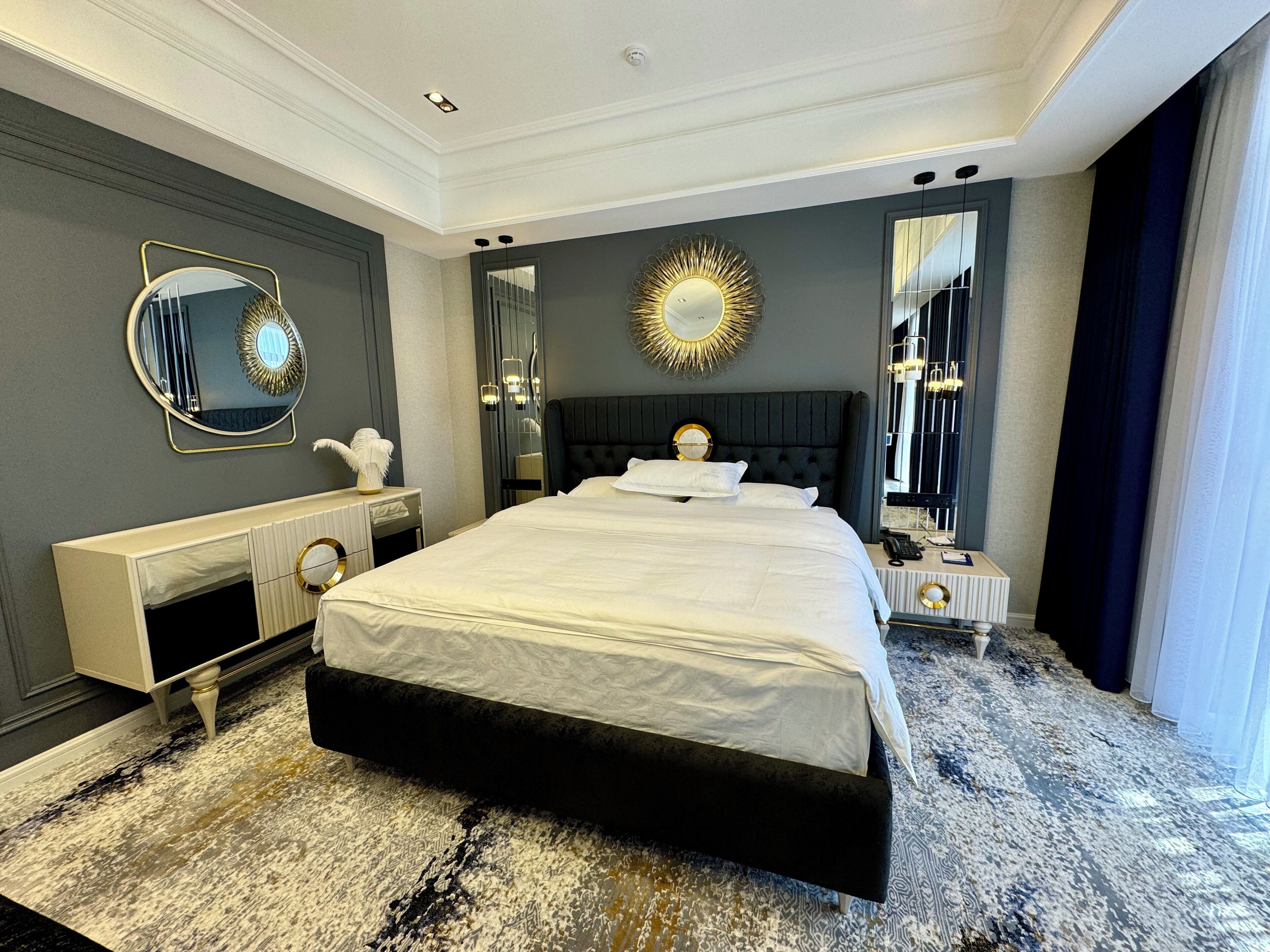 A modern hotel room with a large bed, a vanity with a mirror, and a large window with curtains.
