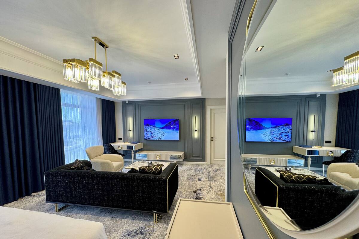 A modern hotel room with a large bed, a seating area with a sofa and armchairs, a TV, and a large mirror.