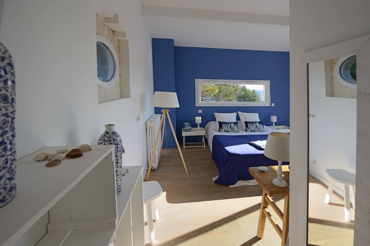 A bedroom with a blue accent wall, a bed with white and blue bedding, a bedside table with a lamp, and a window with a view of trees.