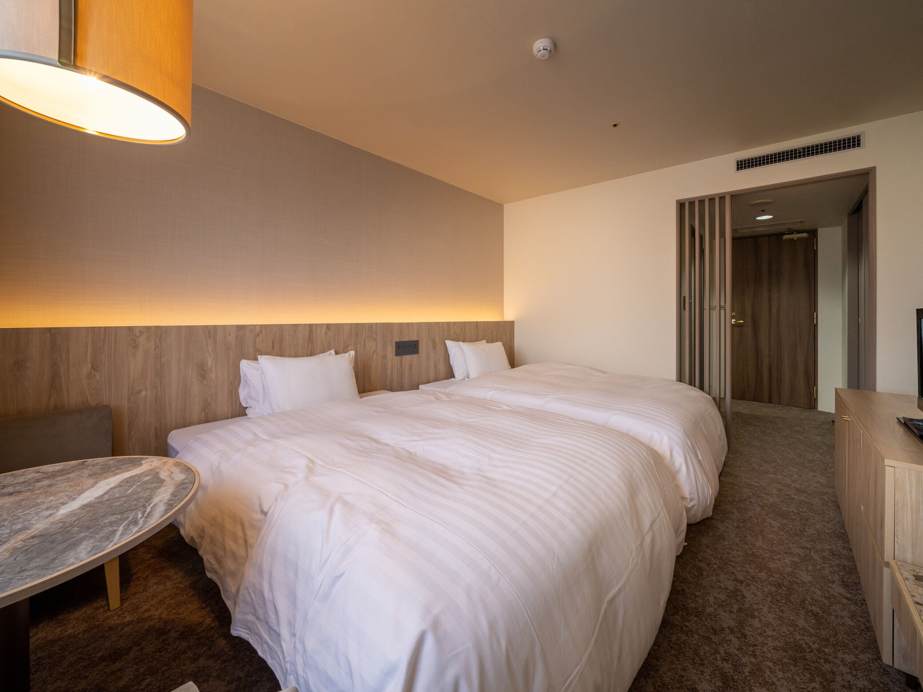 A hotel room with a large bed, a desk, and a chair. There is a wooden headboard and a wall-mounted lamp.