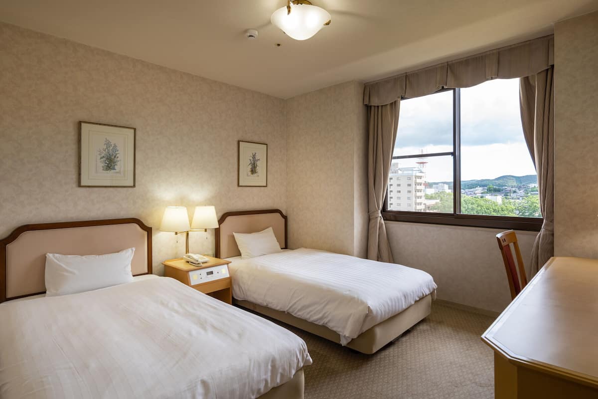 Ein Hotelzimmer mit zwei Betten, einem Schreibtisch, einem Stuhl, einem Nachttisch mit Lampe und einem Fenster mit Blick auf Gebäude und Bäume.