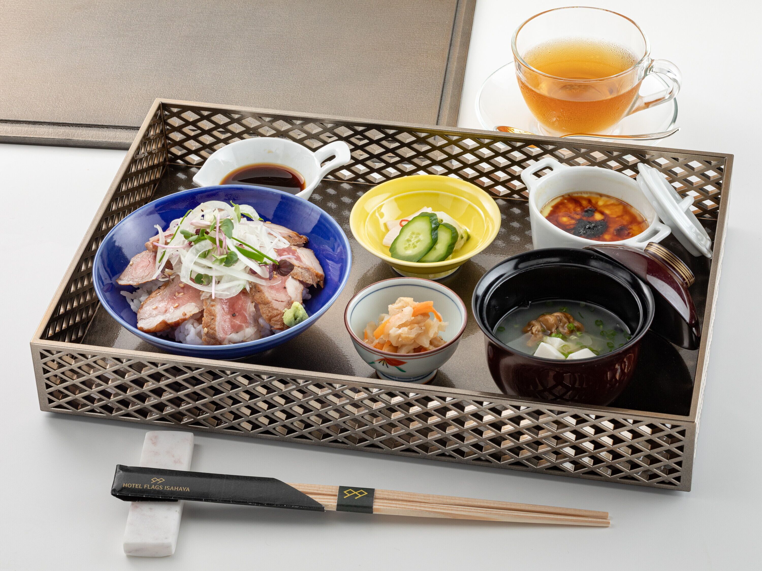 A tray with various Japanese dishes, including a bowl of rice topped with meat and vegetables, a bowl of soup, and a cup of tea.