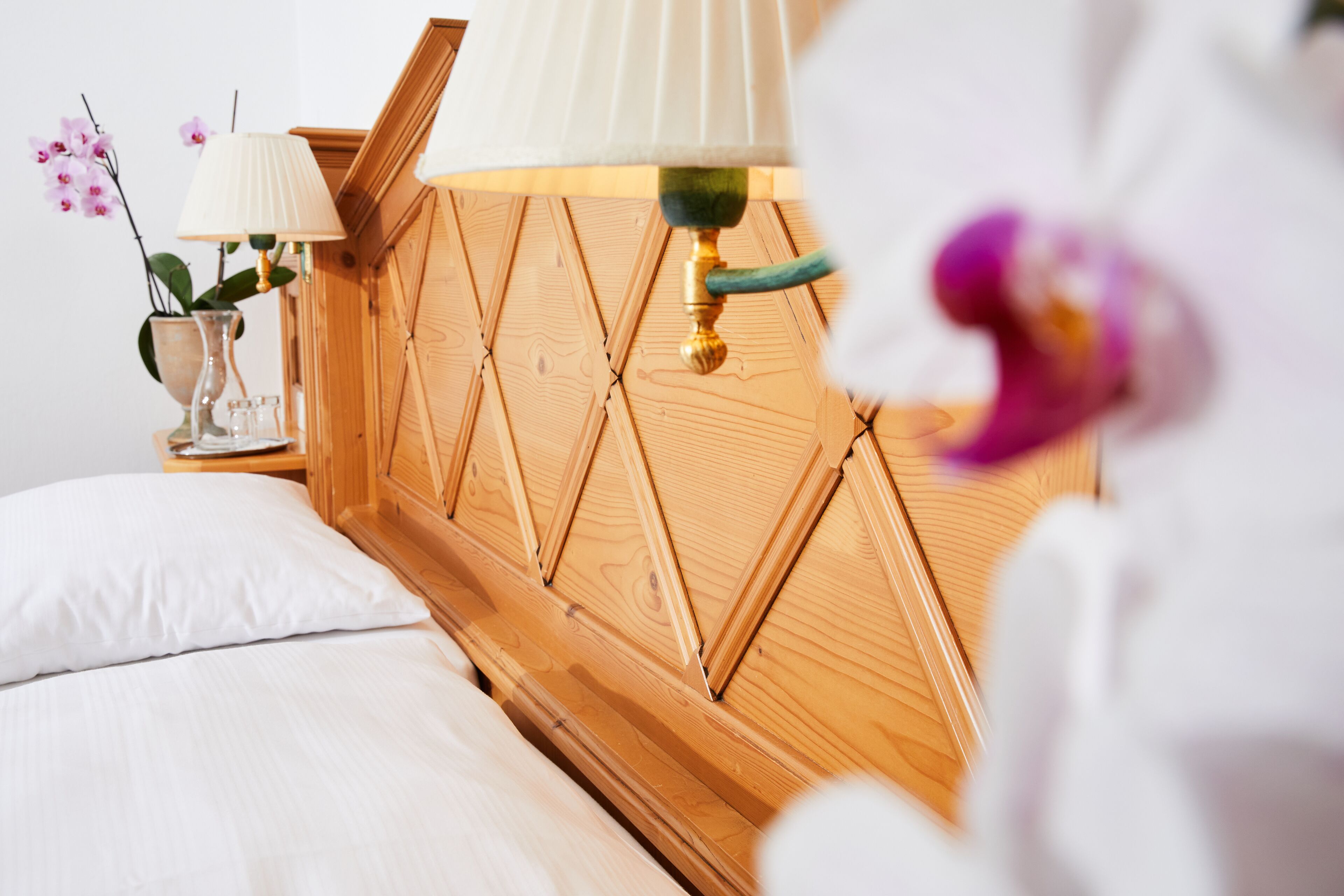 Una cama bien hecha con sábanas blancas, cabecera de madera, mesita de noche con lámpara y un jarrón con flores.