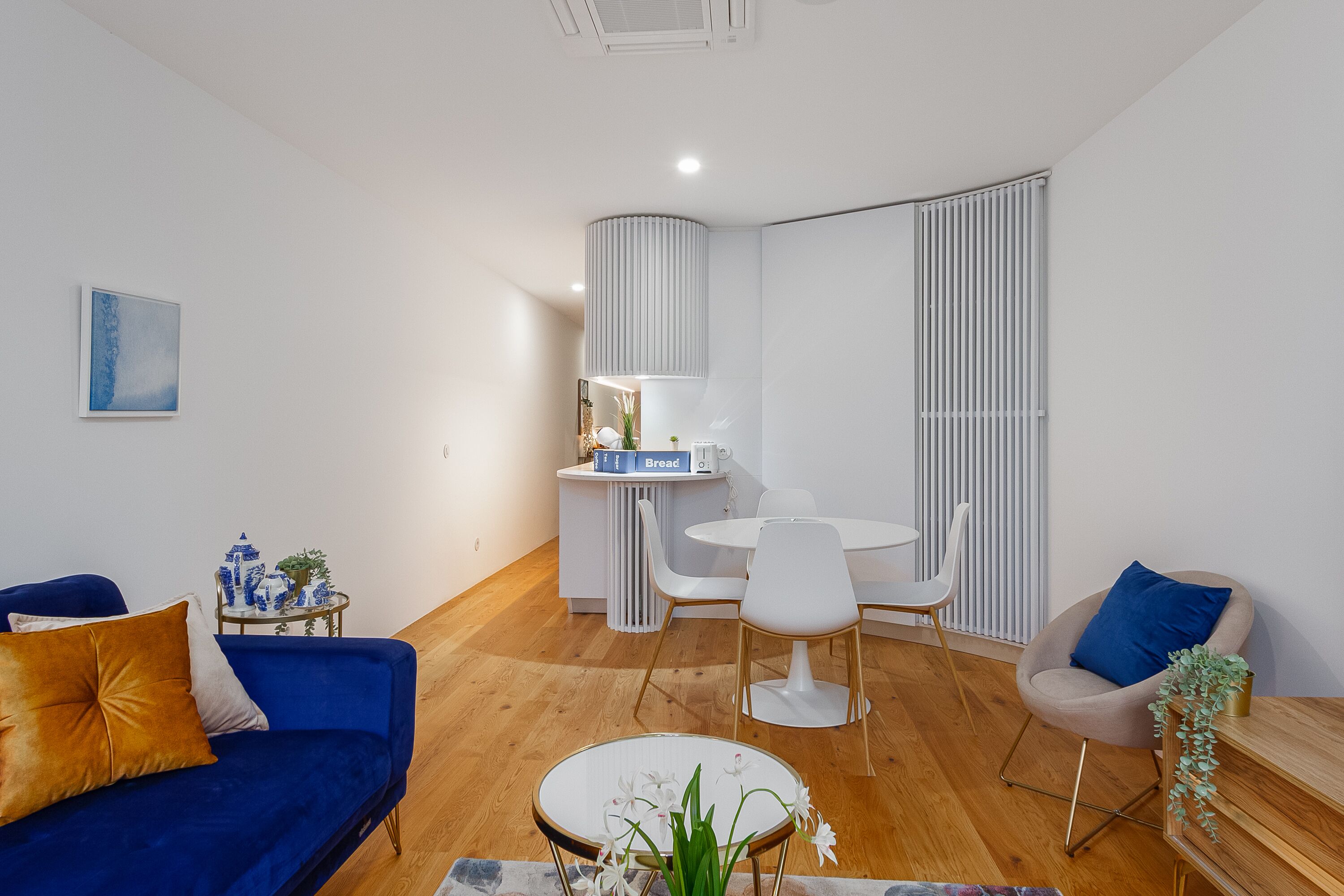 A modern living room with a blue sofa, a round coffee table with flowers, a white dining table with chairs, and a kitchen area with a counter and cabinets.