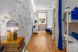 Habitación con suelo de madera, pared blanca con textura, cama con ropa de cama azul, mesita de noche con lámpara y ventana con vistas al exterior.