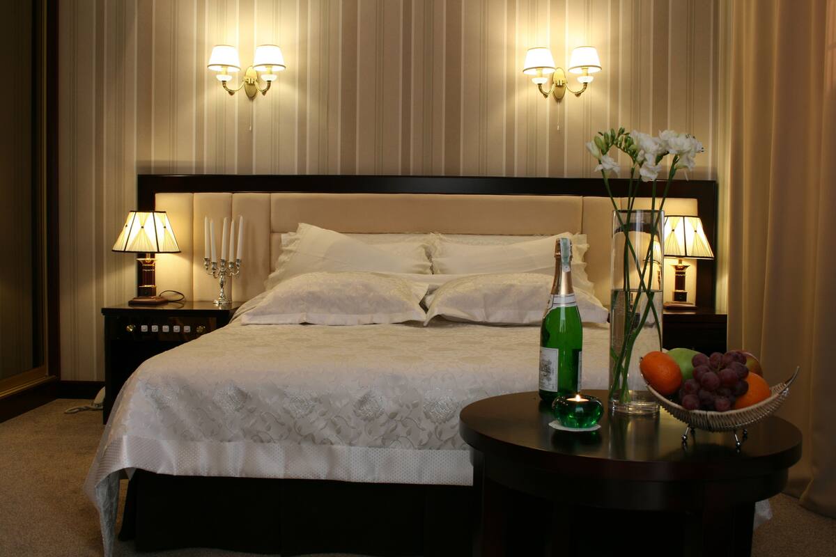 A neatly made bed with white linens, a bedside table with a lamp, a bottle of champagne, and a fruit bowl.