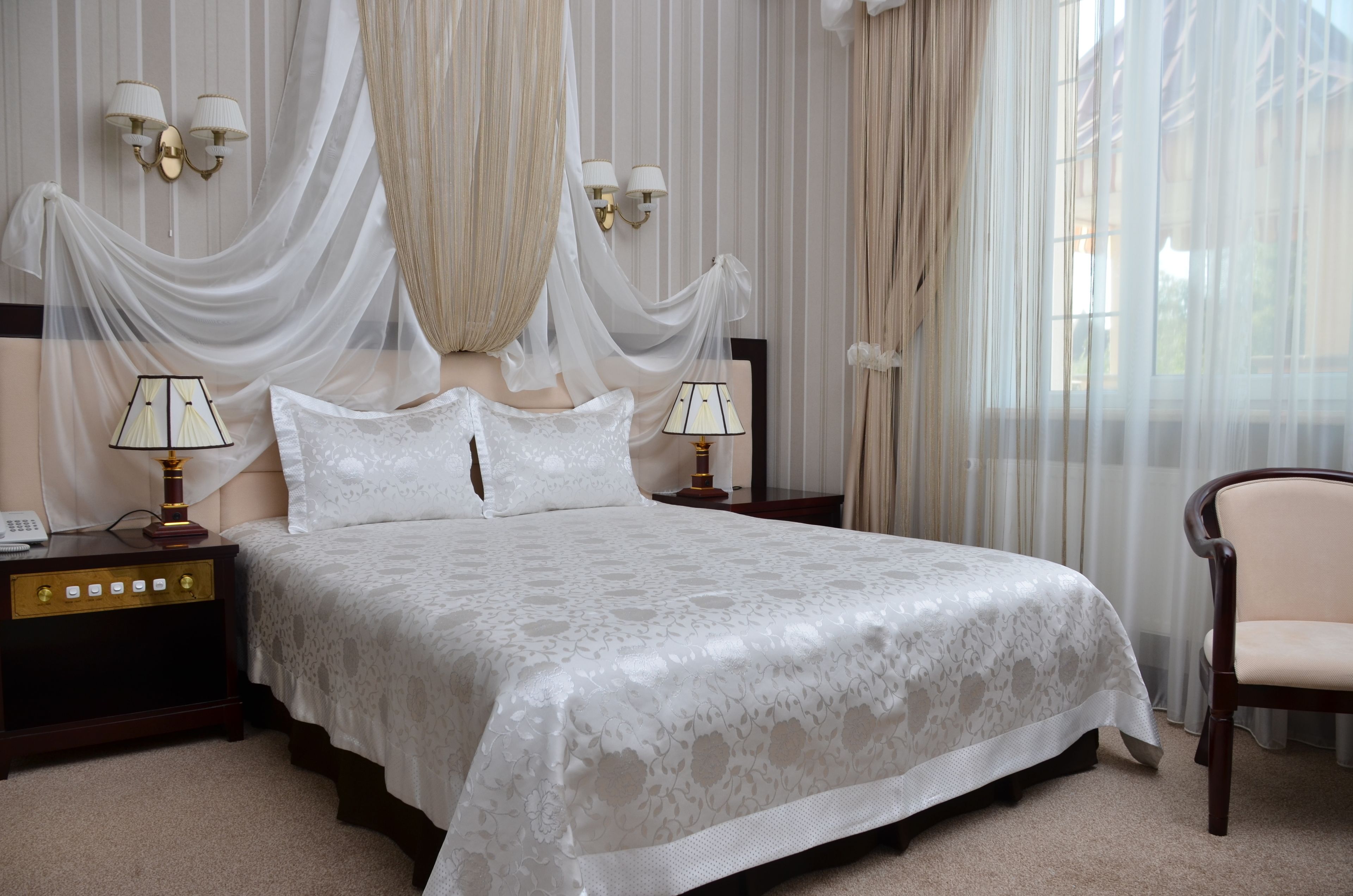 A neatly made bed with a white bedspread and two bedside lamps. There is a chair and a nightstand with a lamp and a phone.