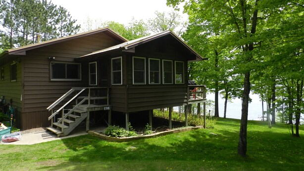 A two-story cabin with a deck, surrounded by lush greenery and a lake.