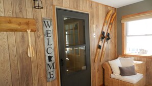 A wooden interior with a 'WELCOME' sign, a door, a wicker sofa, and skis leaning against the wall.