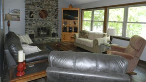 A living room with a stone fireplace, leather furniture, a wooden cabinet with a TV, and large windows.