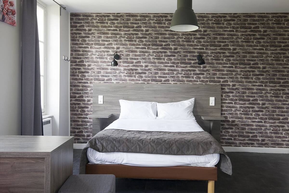 A modern bedroom with a brick-patterned wall, a wooden headboard, a bed with white and gray bedding, and a pendant light.