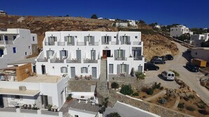 A white building with multiple balconies and a central entrance. There are several cars parked in front, and a white van is also visible. - Hotel Anna (Mykonos)
