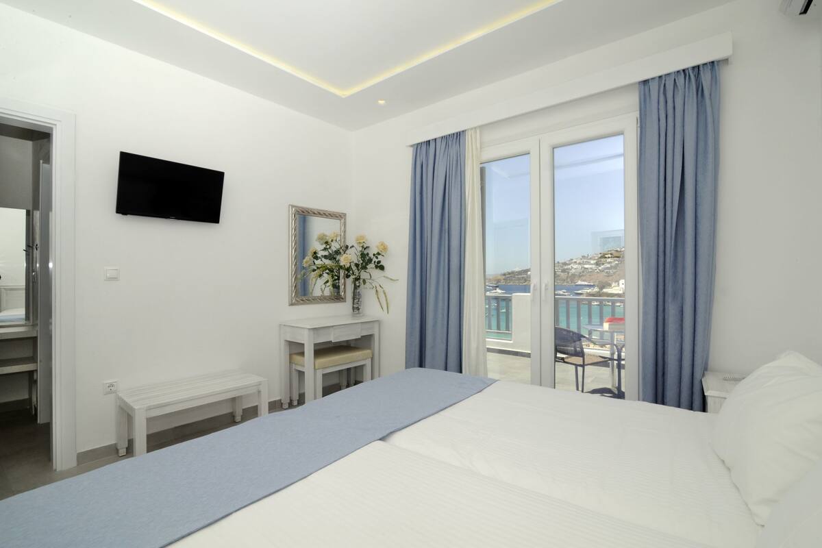 Habitación de hotel con cama, televisión, escritorio con un jarrón de flores y balcón con vistas al mar.
