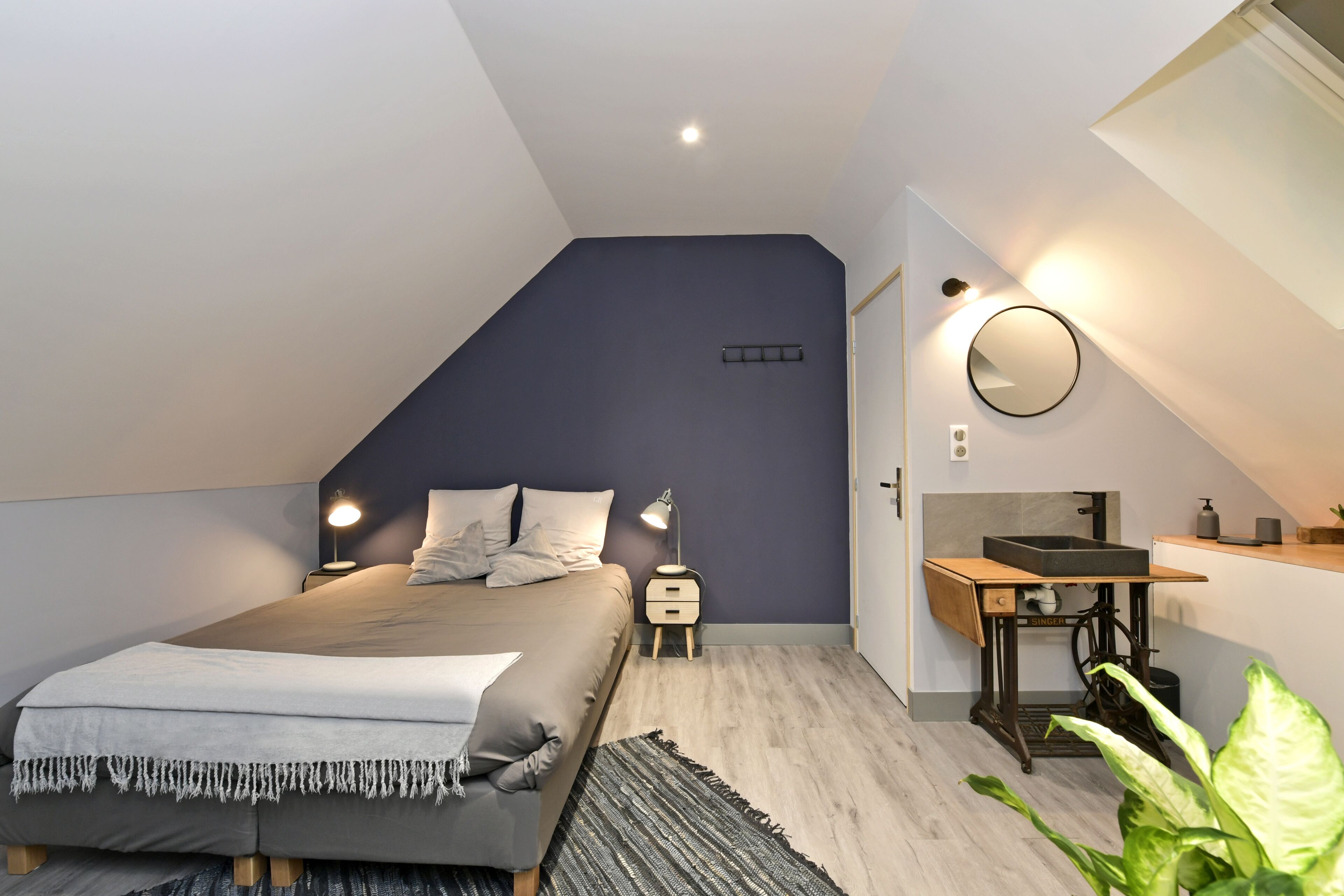 Ein Schlafzimmer mit Bett, Nachttischen, einem Spiegel, einem kleinen Waschbecken und einer Pflanze.