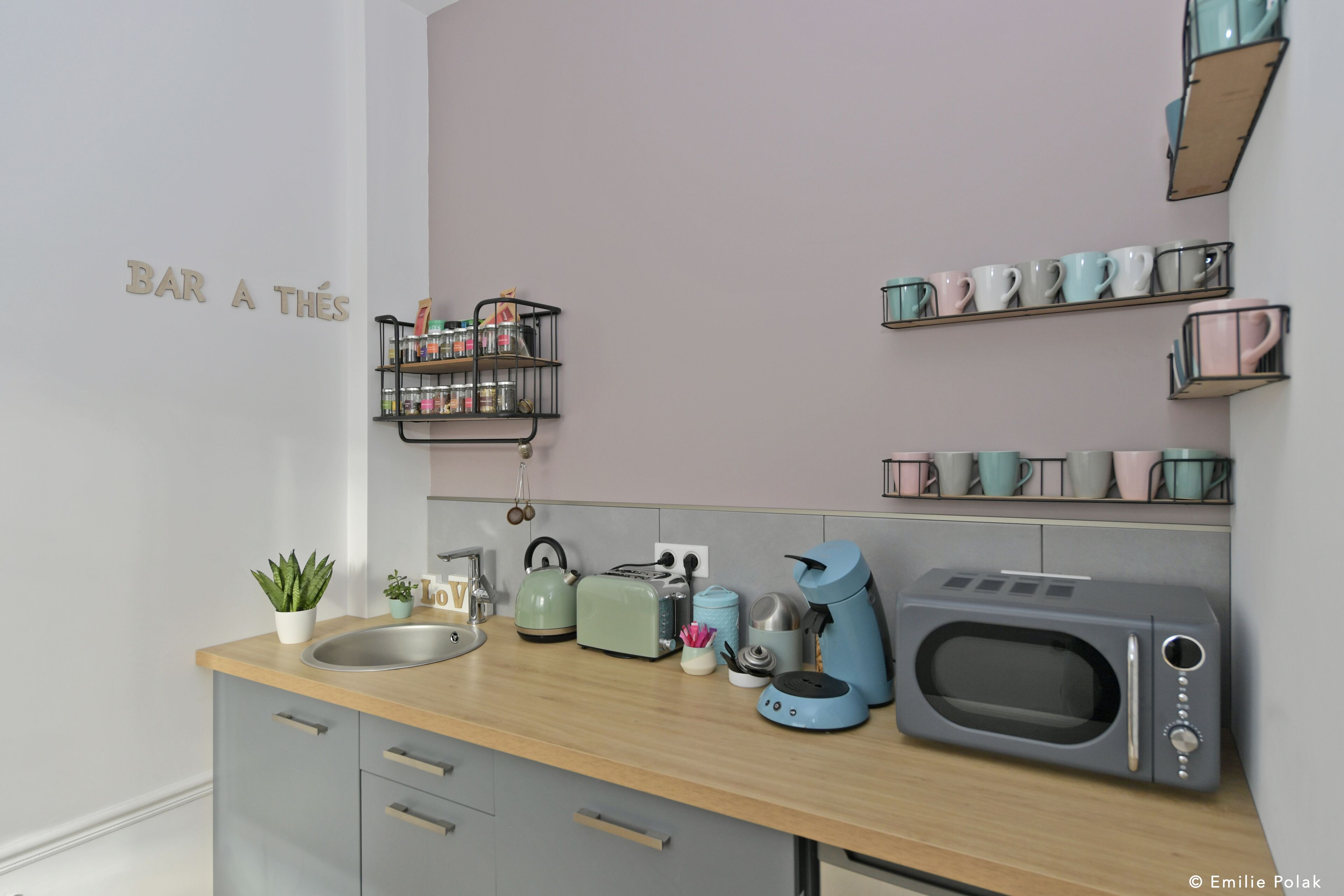 A kitchen with a wooden countertop, a sink, a toaster, a kettle, a coffee maker, and a microwave. There are shelves with mugs and a small plant on the countertop.