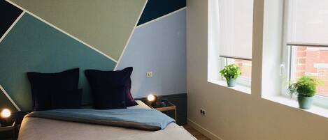 Une chambre moderne avec un motif géométrique sur le mur, un lit recouvert d’une couverture bleue et grise, deux oreillers foncés, des lampes de chevet et une fenêtre donnant sur un bâtiment en briques.