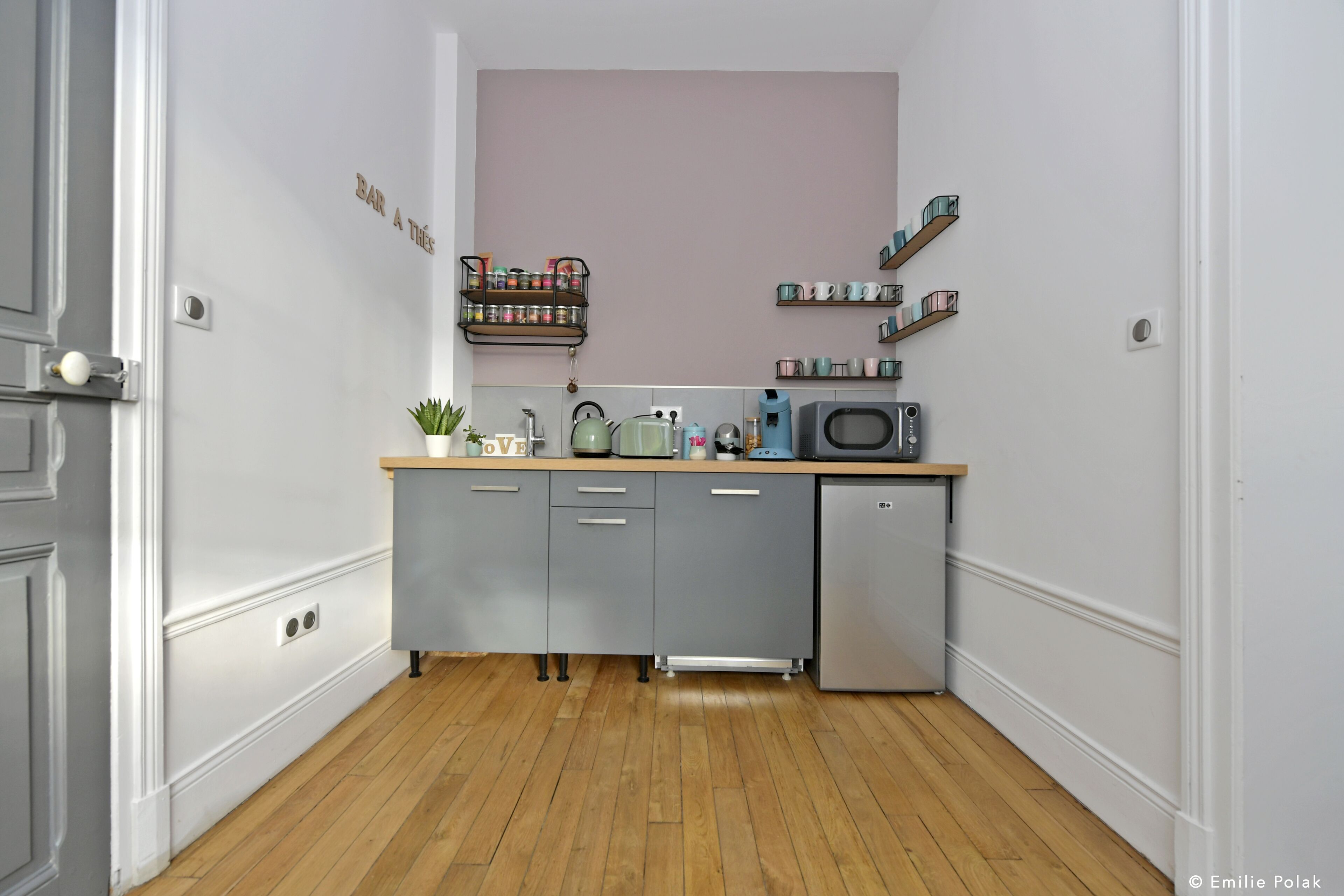 A compact kitchen with grey cabinets, a small fridge, a microwave, and a shelf with jars and cups.