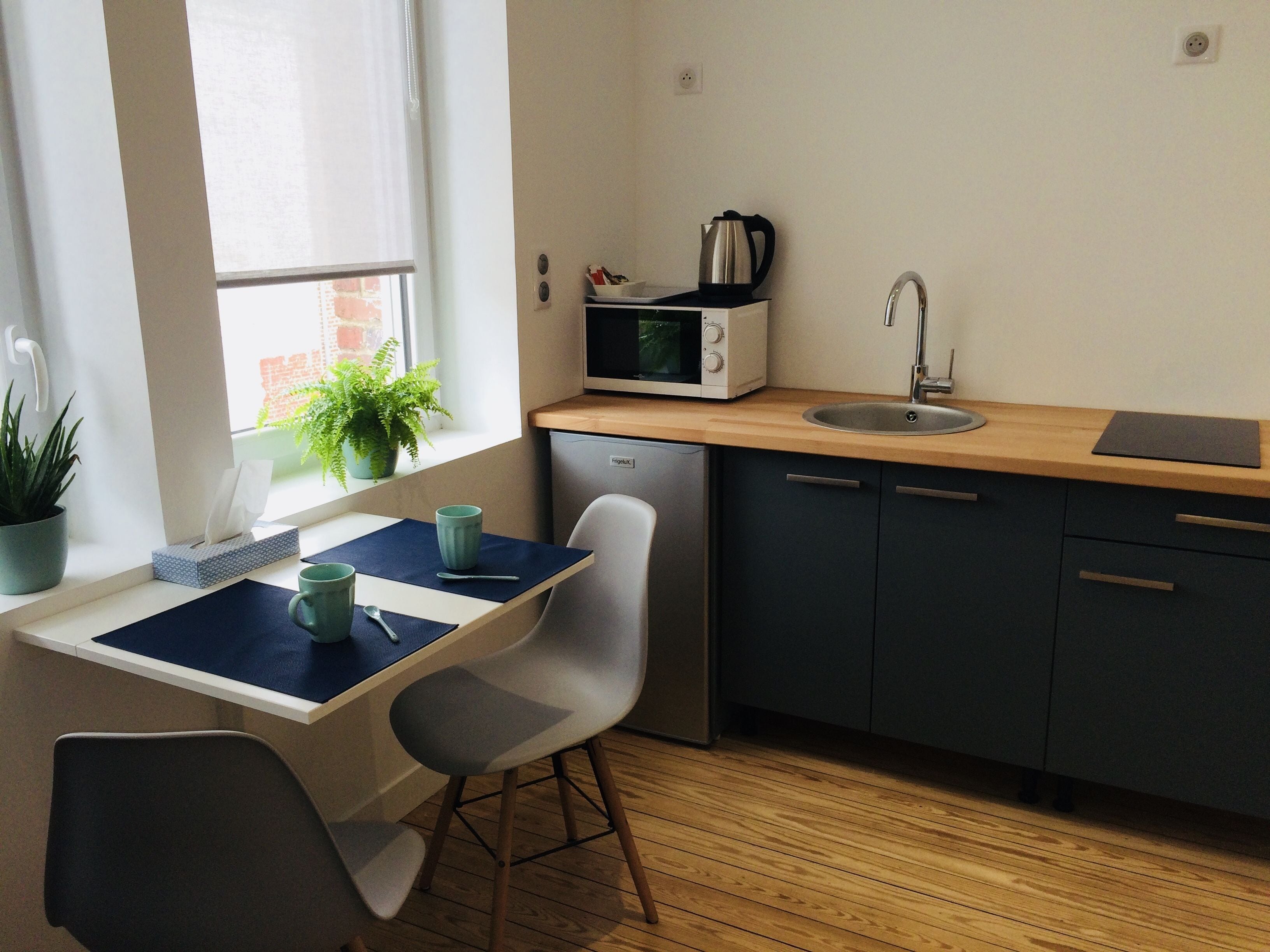 A compact kitchen with a microwave, sink, and a small refrigerator. There is a window with a plant on the windowsill, and a chair is placed at a small table.