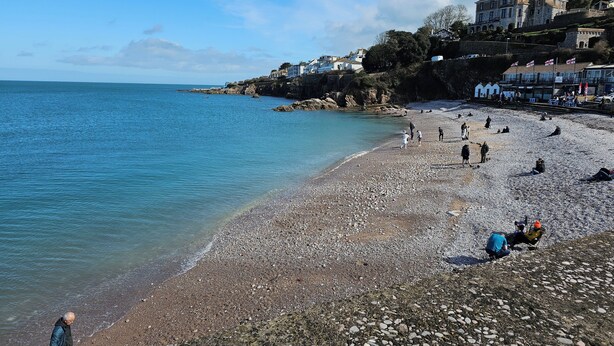 A pebbled beach with people walking and sitting, overlooking a calm sea and a hillside with buildings. - Devoncoast Seaview Apartments (Brixham)