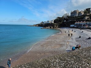 A pebbled beach with people walking and sitting, overlooking a calm sea and a hillside with buildings.