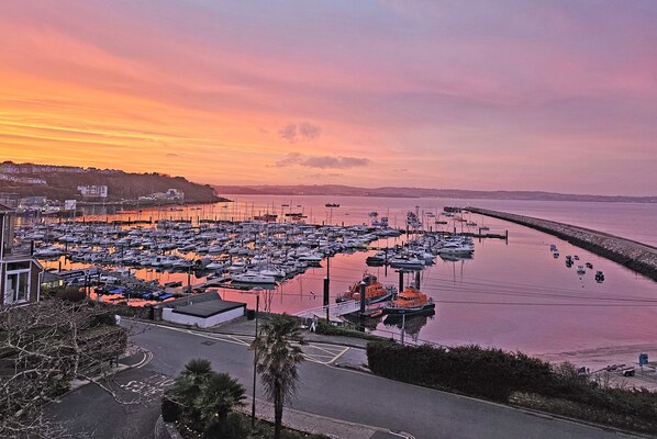 A marina with numerous boats docked, a coastal road, and a backdrop of a sunset sky.