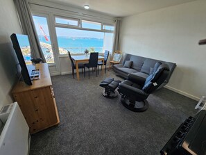 A room with a TV on a wooden stand, a sofa, a dining table with chairs, and a view of the sea through large windows. - Devoncoast Seaview Apartments (Brixham)