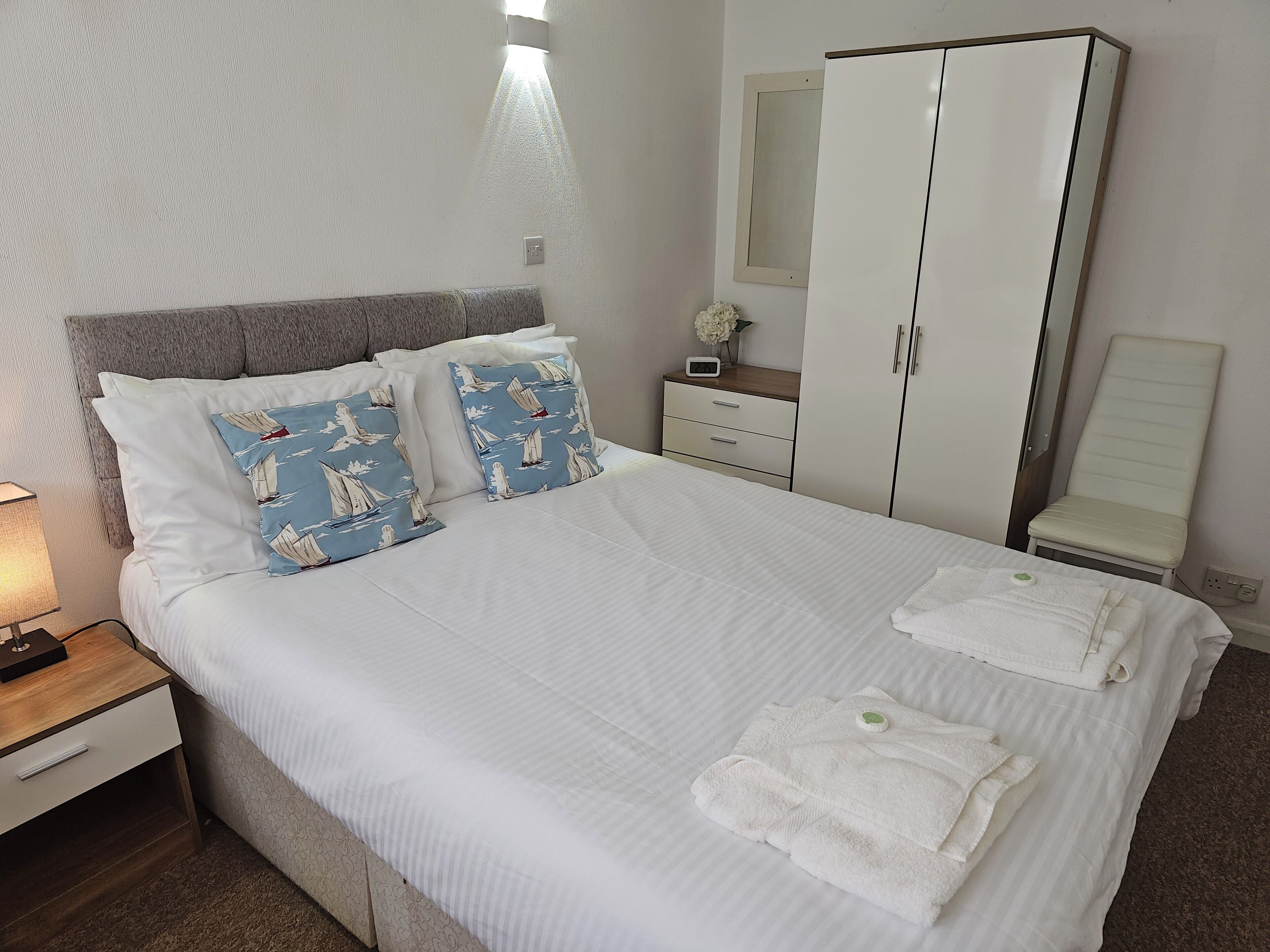A neatly made bed with white linens, two pillows with a sailboat pattern, a bedside table with a lamp, a nightstand, a wardrobe, and a chair.