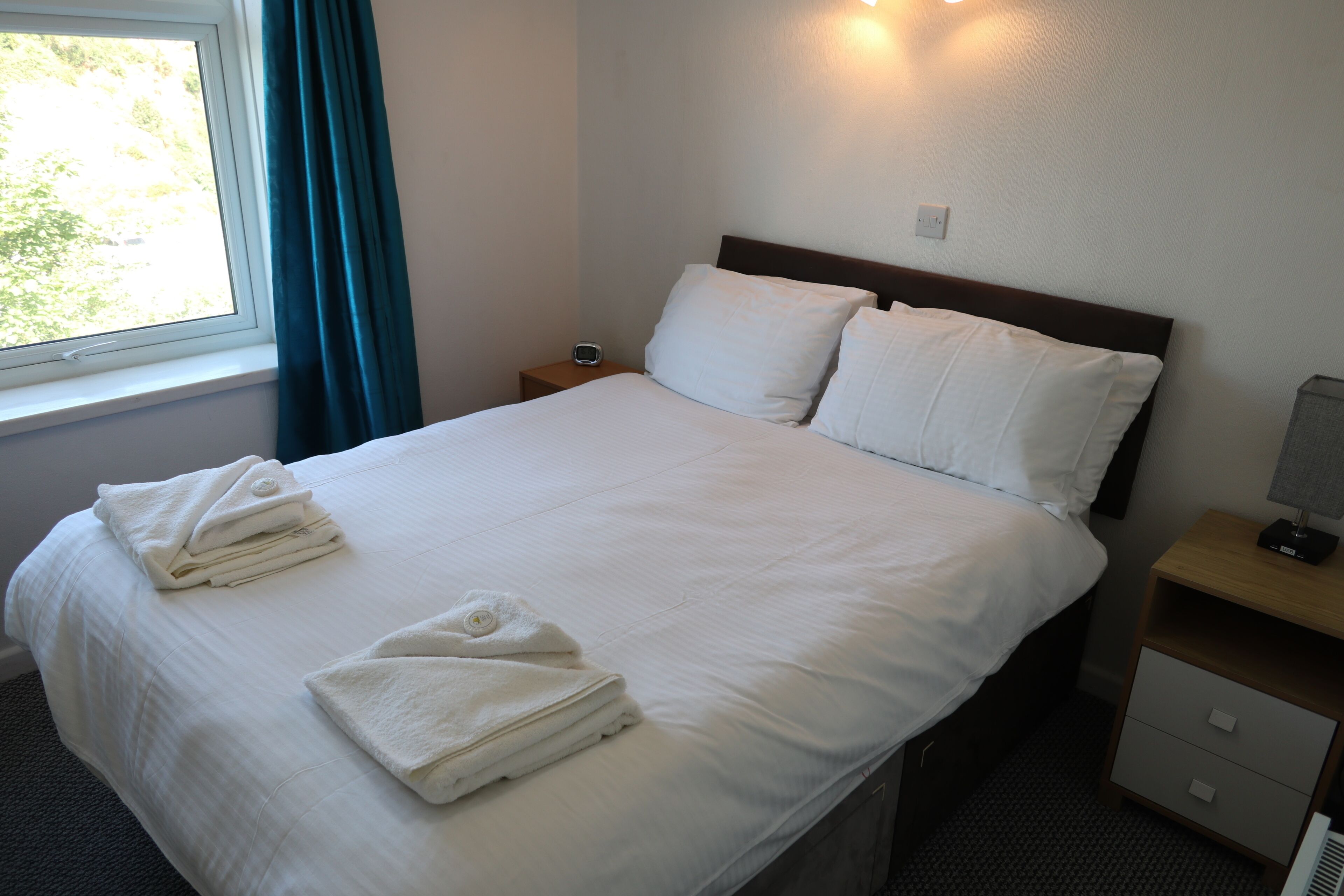 A neatly made bed with white linens and towels, a bedside table with a lamp, and a window with curtains.