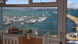 A view from a room with a large window overlooking a marina with numerous boats, a lighthouse, and a rocky breakwater. - Devoncoast Seaview Apartments (Brixham)
