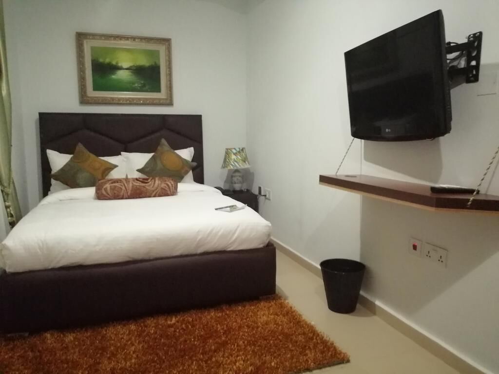 A bedroom with a bed, a mounted TV, a painting on the wall, and a small trash bin.