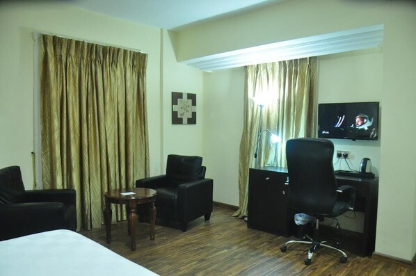 Habitación de hotel con una cama, dos sillones negros, una mesita de madera, un escritorio con una computadora y un televisor montado en la pared.