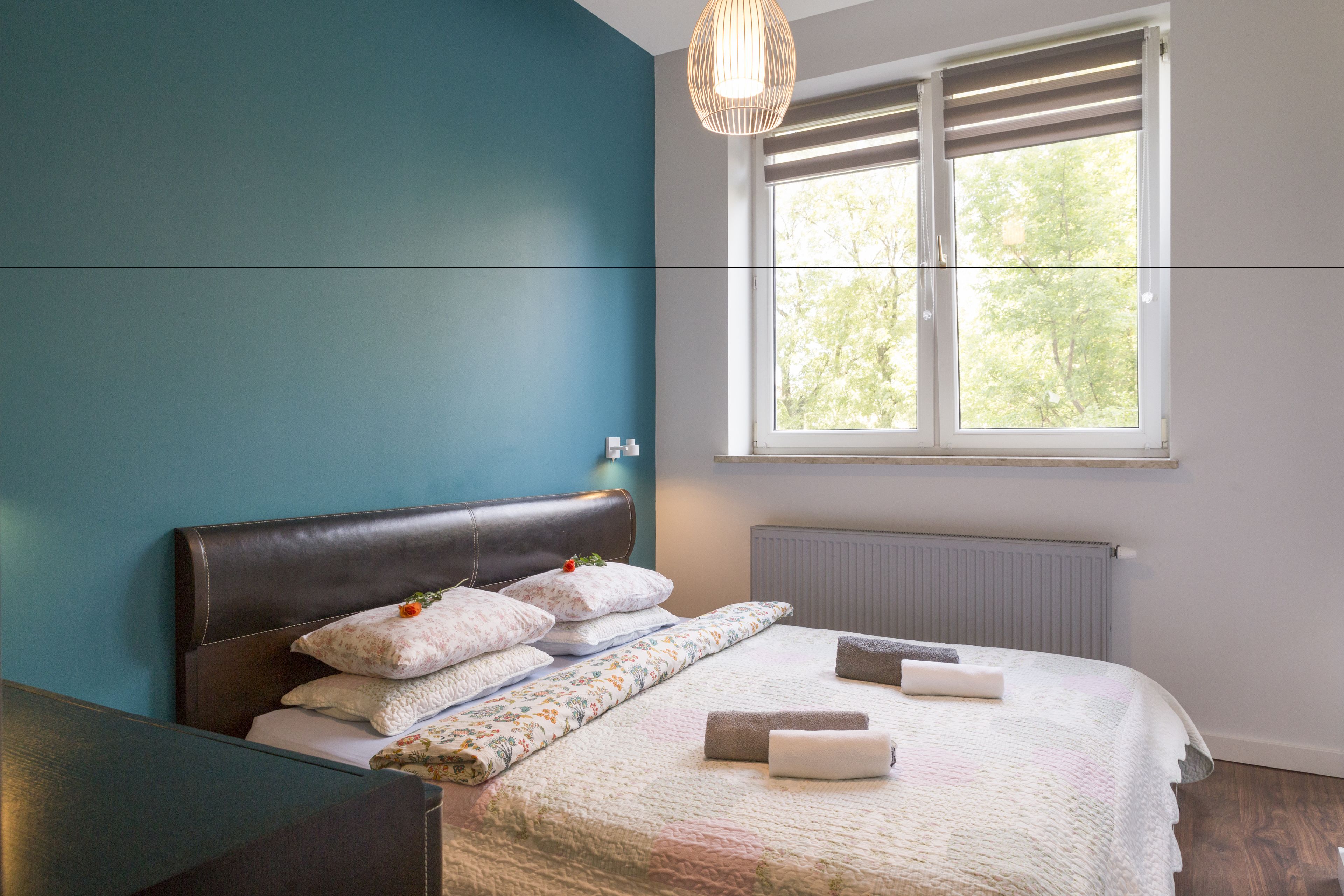 Un dormitorio moderno con una pared de acento color turquesa, un ventanal con persianas, un cabecero marrón oscuro y toallas ordenadas.