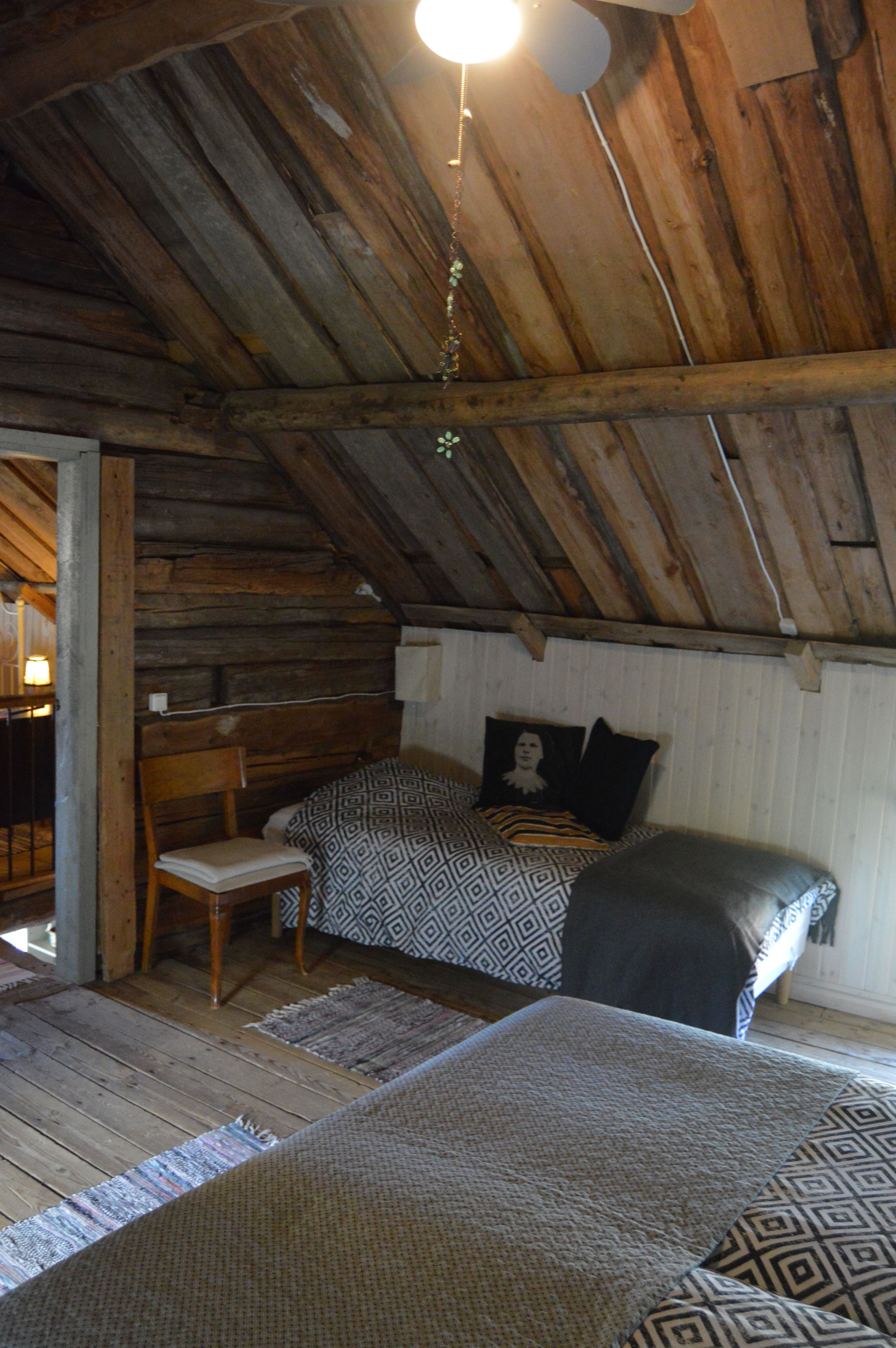 A rustic room with a sloped ceiling, wooden beams, a bed with patterned bedding, a chair, and a small table.