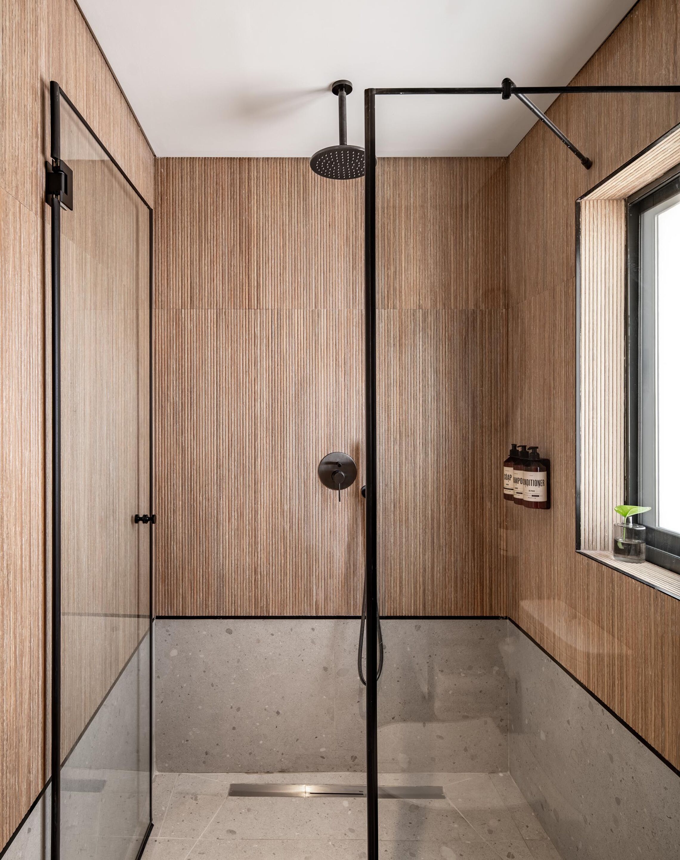 A modern shower with a glass door, wooden panel walls, and a window with a plant.