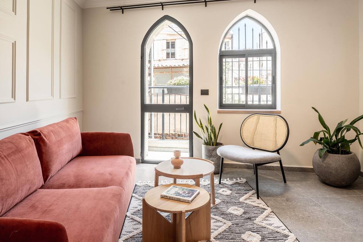 A room with a red sofa, a wooden coffee table, a wicker chair, and two windows with arched tops.